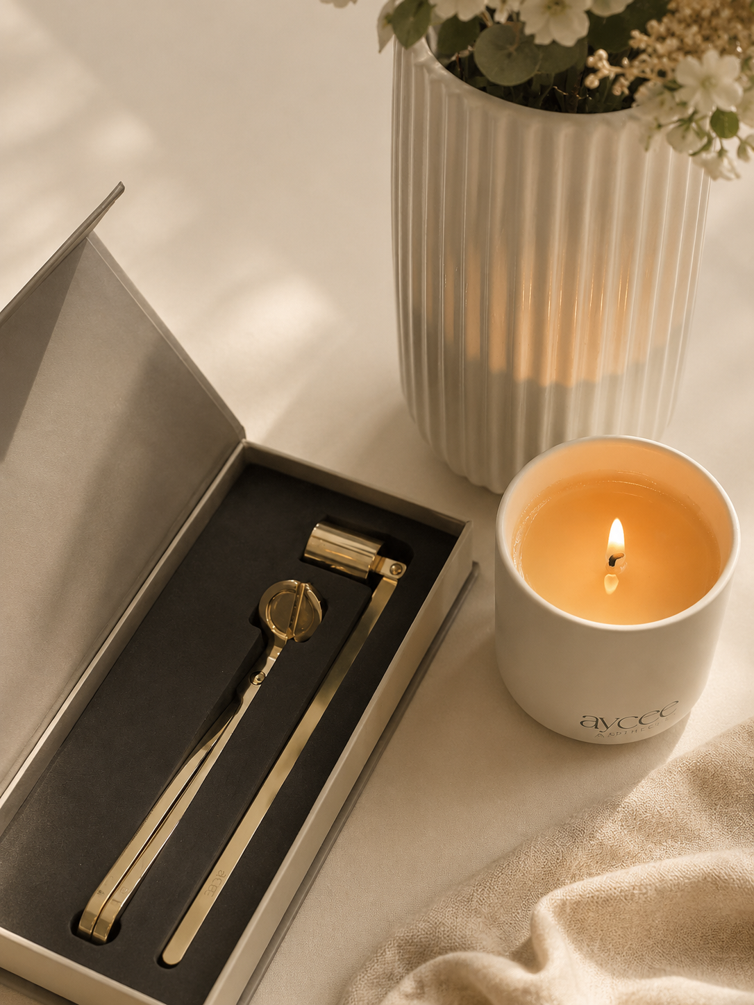 Gold candle care tools in a box next to a lit ceramic candle jar and vase