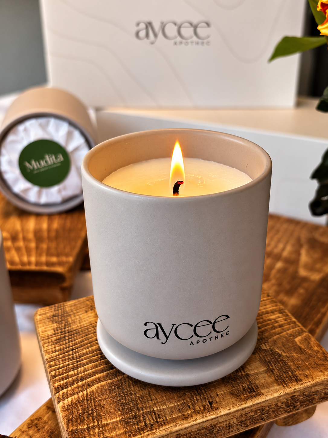 A lit Aycee Apothec candle in a matte light gray ceramic jar, resting on a rustic wooden block. The background shows the product's white packaging box and another candle tin.