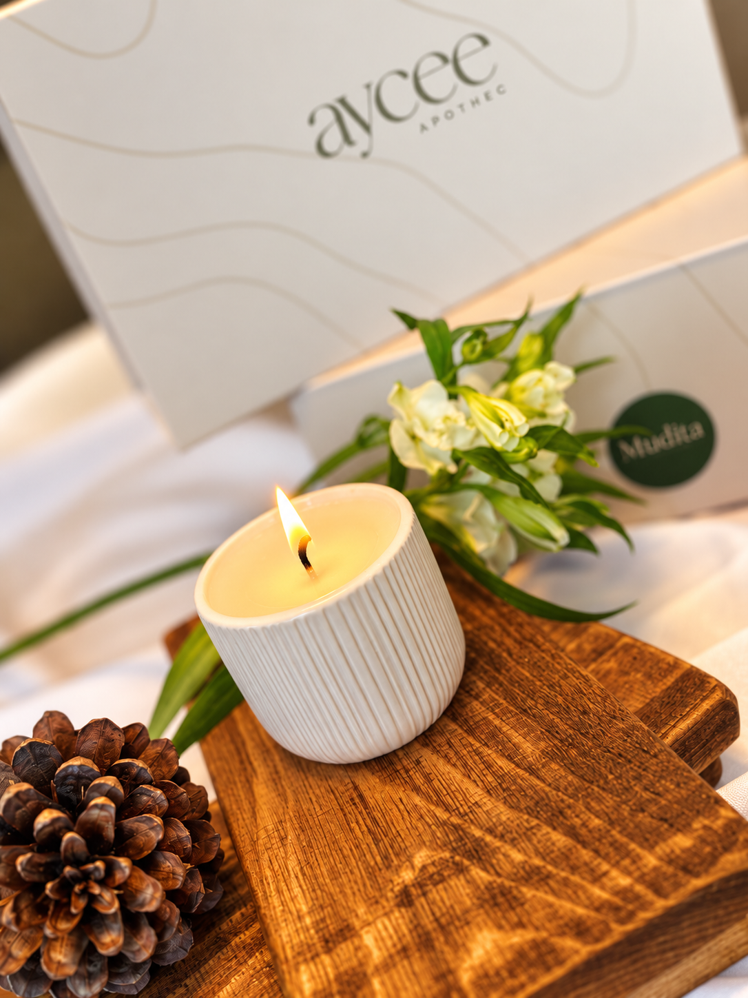 A lit white ribbed candle holder candle sits on a wooden board with a pinecone and white flowers in the foreground, with Aycee Apothec branded gift boxes in the background.