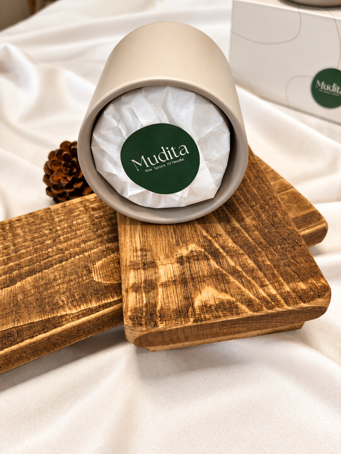 A ceramic cylindrical container with a lid, shown open and tipped on its side on a wooden block. The inside is lined with white tissue paper and features a green circular label reading 'Mudita'.