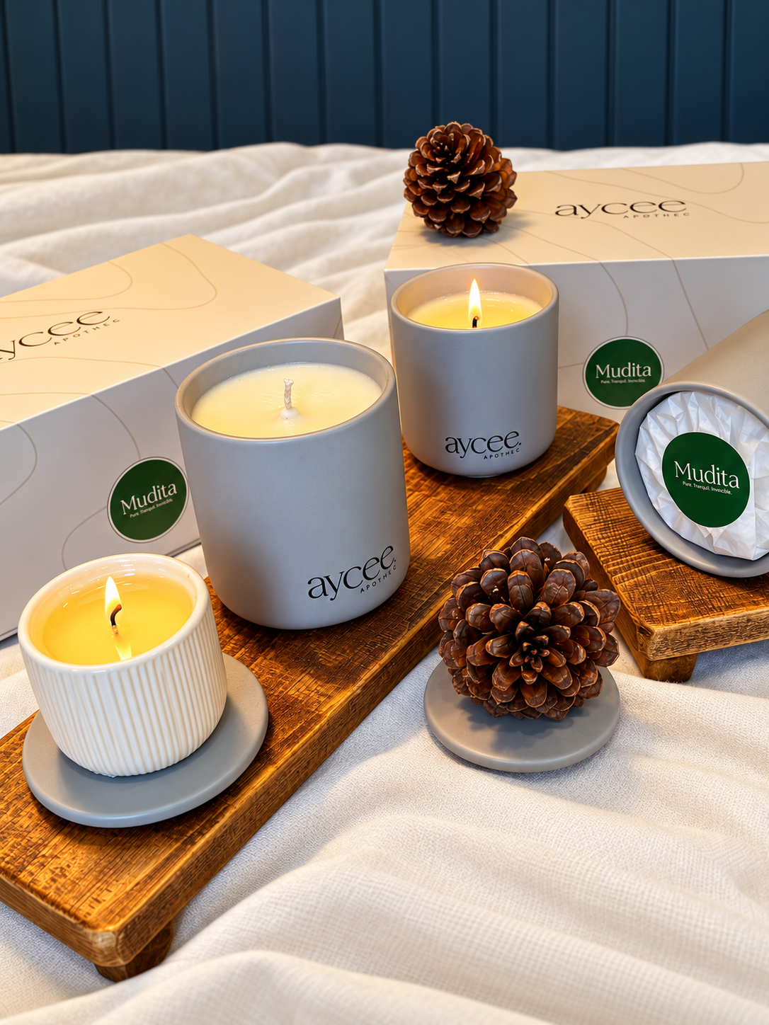 A styled product display of aycee candles by Mudita, featuring three different candle vessels (a large matte gray jar, a small ribbed white cup, and a round tin) arranged on a wooden tray with pinecones on a white fabric background.