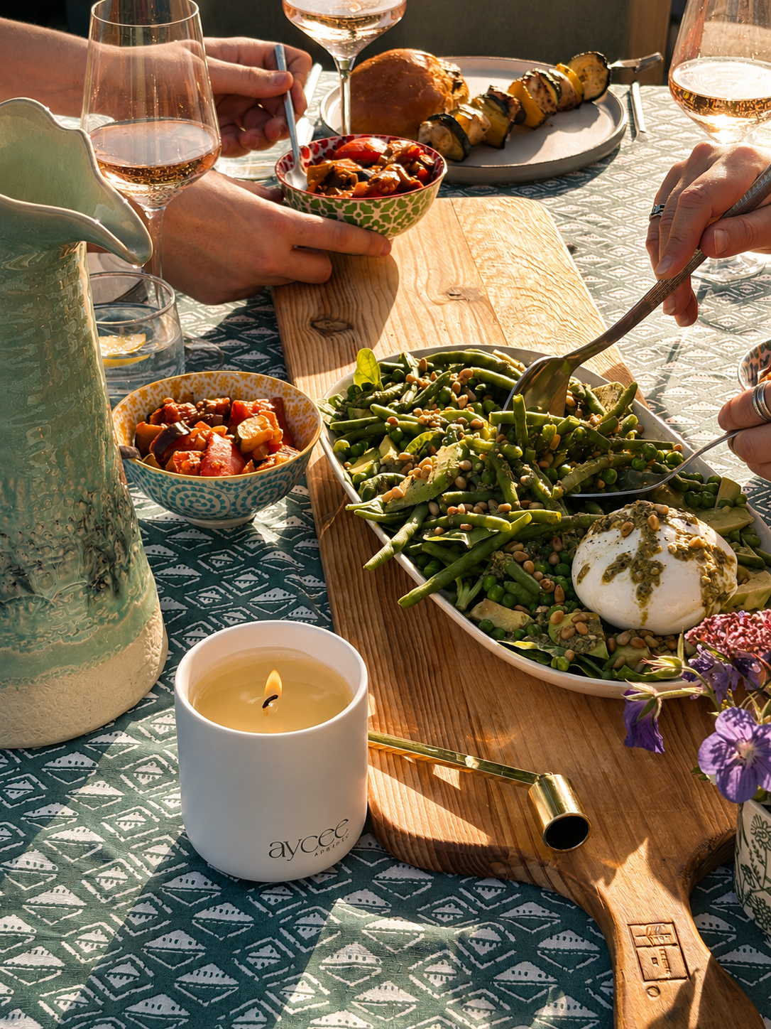 People enjoying a meal outdoors with a candle and cutlery on a patterned tablecloth.