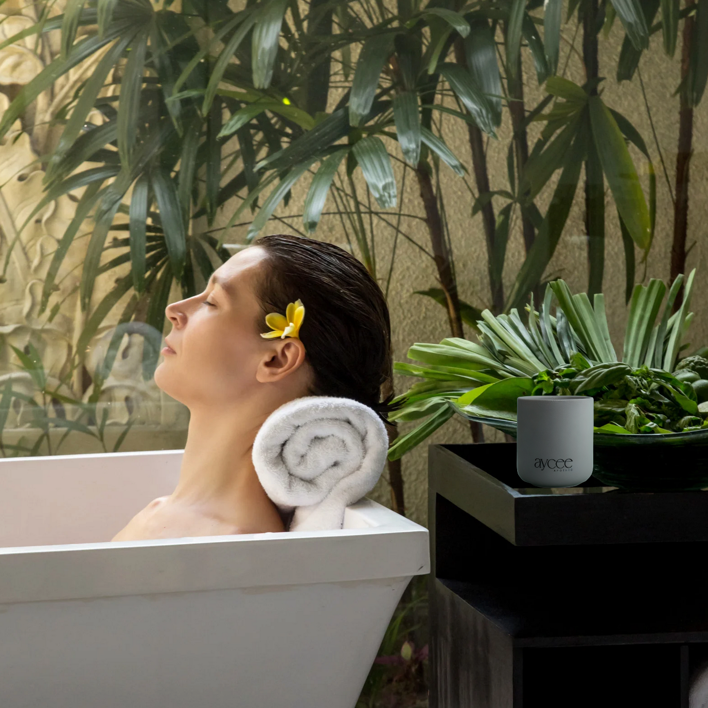 Woman relaxing in bath with greenery around er and an Aycee Apothec candle on a side table